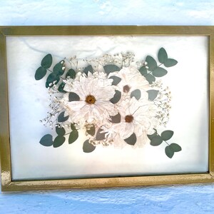 May include: A framed floral arrangement featuring white flowers with brown centers, surrounded by small white blossoms and green leaves. The arrangement is encased in a gold-colored rectangular frame, creating a decorative piece.