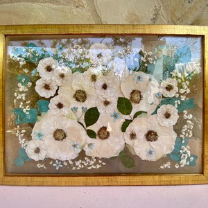 May include: A framed pressed flower art piece featuring white flowers with blue accents. The flowers are arranged in a cluster and are surrounded by greenery. The frame is gold and has a glass front.