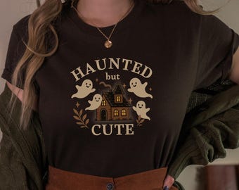 May include: A dark brown t-shirt with a Halloween-themed graphic. The design features the words "HAUNTED but CUTE" with a haunted house, ghosts, and decorative elements. The shirt is a short-sleeved crew neck.