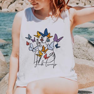 May include: White tank top featuring a line art design of a face and butterflies in shades of blue, yellow, and purple. The text "Fuck Trump" is written in black script below the design.