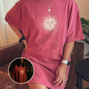 Könnte beinhalten: Ein kurzärmeliges T-Shirt mit Rundhalsausschnitt in verwaschenem Rot. Auf der Vorderseite befindet sich ein Grafikdruck einer Sonne mit menschlichem Gesicht, umgeben von Strahlen, hängenden Sternen und einer Mondsichel. Ein kleiner runder Ausschnitt zeigt das gleiche T-Shirt-Design.
