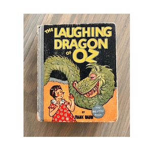 Può includere: Una copertina di libro vintage con un drago verde con un ghigno pieno di denti, una bambina con un vestito a pois rosso e bianco, e il titolo "The Laughing Dragon of Oz" di Frank Baum. Il libro fa parte della serie "The Big Little Book".