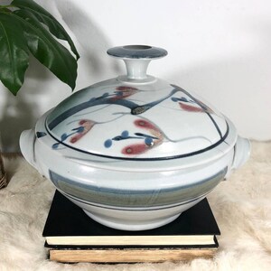 May include: A white ceramic serving dish with a lid. The dish has a blue and gray design with red and blue floral accents. The lid has a small knob on top.