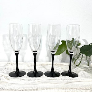 May include: Four clear glass champagne flutes with black stems. The flutes are arranged in a row on a white crocheted surface.