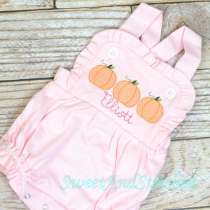 May include: A light pink baby romper featuring a ruffled neckline and two white buttons. The chest area displays an embroidered design of three orange pumpkins with green stems and the name Elliott stitched in pink cursive lettering.
