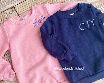 Op de afbeelding: Twee gepersonaliseerde sweatshirts, één roze met de naam "Ava" geborduurd in paars en één donkerblauw met de naam "CJY" geborduurd in wit. De sweatshirts liggen op een houten oppervlak.