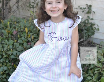 May include: A young girl wearing a white and pink striped dress with a purple embroidered name "Ava" on the front. The dress has a ruffled hem and cap sleeves. The girl is smiling and standing in front of a green bush.
