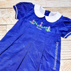 May include: A blue corduroy dress with white collar and short sleeves. The dress has a white embroidered design of a ballerina, a snowman, and a reindeer with the text "Winter Girl" and "briar".