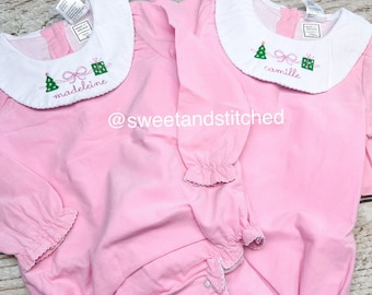 May include: Two pink long-sleeved baby onesies with white Peter Pan collars and green embroidered Christmas trees and presents. The onesies have the names "madeleine" and "camille" embroidered on them. The onesies are on a wooden surface. @sweetandstitched