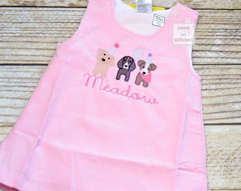 May include: Pink sleeveless dress with embroidered dogs wearing party hats and the name "Meadow" on the front. The dress has a white undershirt and a small tag that says "sweet and stitched".