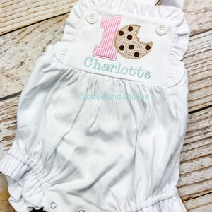 K&ouml;nnte beinhalten: Wei&szlig;er Baby-Strampler mit pink-wei&szlig; gestreiftem Nummer eins und einem braunen Schokoladenkeks mit einem Bissen heraus. Der Strampler ist personalisiert mit dem Namen "Charlotte" und dem Text "@sweetandstitched".