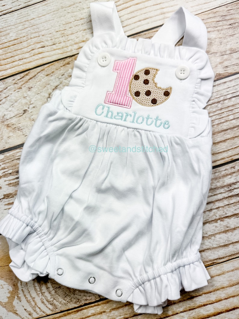 K&ouml;nnte beinhalten: Wei&szlig;er Baby-Strampler mit pink-wei&szlig; gestreiftem Nummer eins und einem braunen Schokoladenkeks mit einem Bissen heraus. Der Strampler ist personalisiert mit dem Namen "Charlotte" und dem Text "@sweetandstitched".