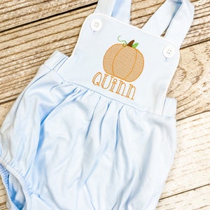 May include: Light blue baby romper with white trim and a pumpkin design. The romper has a name, "quinn", embroidered on the front.
