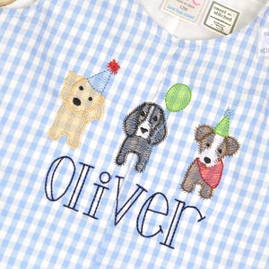 May include: Blue and white gingham fabric baby onesie with embroidered dogs wearing party hats and the name "Oliver" in blue thread.