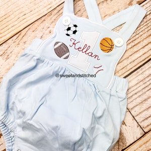 May include: Light blue baby onesie with a white embroidered number one, the name "Kellan" and various sports balls. The onesie has white straps and white buttons. The text "@sweetandstitched" is embroidered on the bottom.
