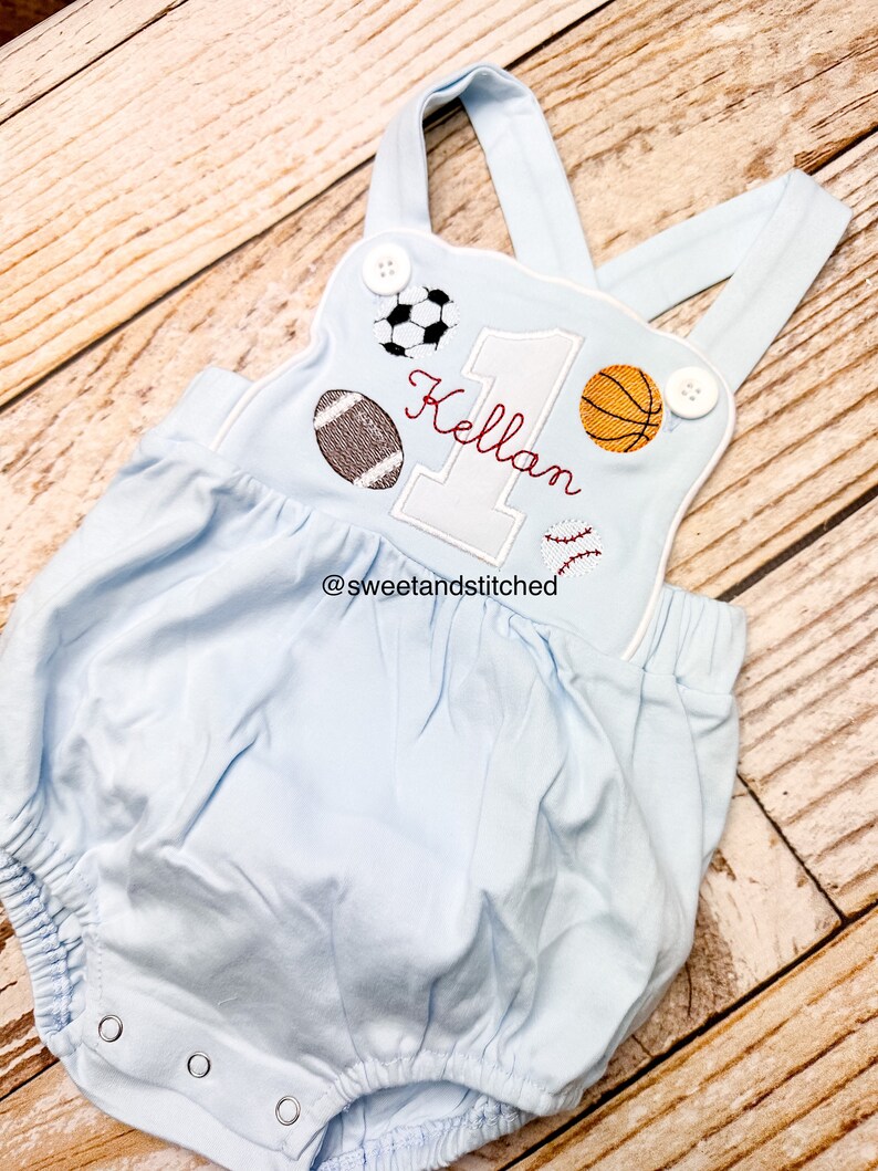 May include: Light blue baby onesie with a white embroidered number one, the name "Kellan" and various sports balls. The onesie has white straps and white buttons. The text "@sweetandstitched" is embroidered on the bottom.