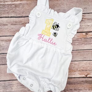 May include: White baby onesie with a yellow number one and two bees embroidered on it. The name Hallie is embroidered below the number one in pink thread.