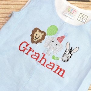 May include: Light blue baby onesie with a red embroidered name "Graham" and animal illustrations including a lion, elephant, and zebra. The onesie has a white label with the text "sweet and stitched".