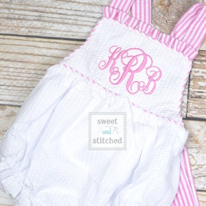 May include: White baby romper with pink and white striped straps and a pink monogram. The romper has a ruffled neckline and a gathered skirt. The text "sweet and stitched" is printed on a tag attached to the romper.