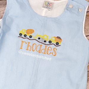 May include: Light blue romper with embroidered construction vehicles, including a cement mixer, excavator, and dump truck. The name "Rhoades" and the text "@sweetandstitched" are stitched in orange thread. The romper has white trim and button closures.