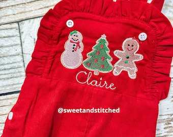 May include: Red corduroy overall romper with a snowman, Christmas tree, and gingerbread man embroidered on the front. The name "Claire" is embroidered below the figures. The text "@sweetandstitched" is embroidered at the bottom of the romper.