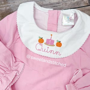 May include: Pink corduroy baby dress with a white scalloped collar. The collar features embroidered pumpkins, a cake, and the name "Quinn". The sleeves have ruffled cuffs. The text "@sweetandstitched" is also embroidered on the dress.