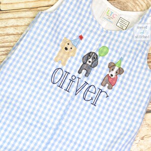 May include: A blue and white gingham baby onesie with the name "Oliver" embroidered on the front. Three embroidered dogs wearing party hats are above the name. The onesie has a tag that says "sweet and stitched".