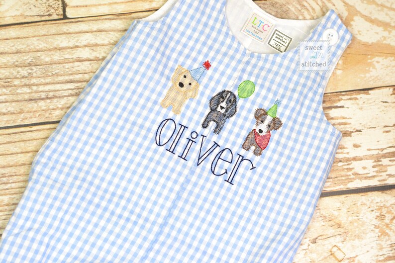 May include: A blue and white gingham baby onesie with the name "Oliver" embroidered on the front. Three embroidered dogs wearing party hats are above the name. The onesie has a tag that says "sweet and stitched".