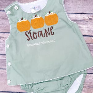 May include: A two-piece baby outfit in a soft green color, featuring an embroidered pumpkin design. The top has three orange pumpkins with green stems and the name "Sloane" embroidered in brown. The outfit has white trim and button closures.
