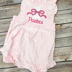May include: Pink baby onesie with a white number one and a pink bow. The onesie has a ruffle trim and says "Parker" on the front.