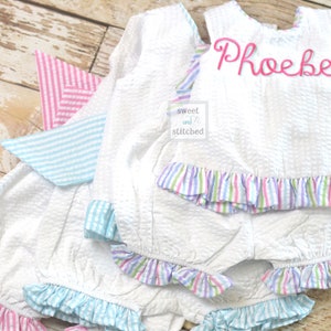 Puede incluir: Conjunto de beb&eacute; de seersucker blanco con ribete de volantes a rayas rosas, azules y moradas. La parte superior tiene el nombre "Phoebe" bordado en rosa. El conjunto incluye una parte superior y un bloomers a juego.