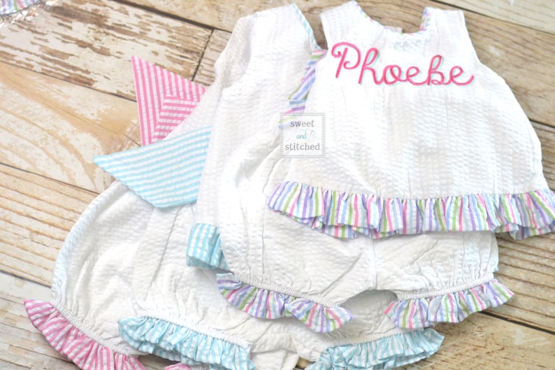 Puede incluir: Conjunto de beb&eacute; de seersucker blanco con ribete de volantes a rayas rosas, azules y moradas. La parte superior tiene el nombre "Phoebe" bordado en rosa. El conjunto incluye una parte superior y un bloomers a juego.