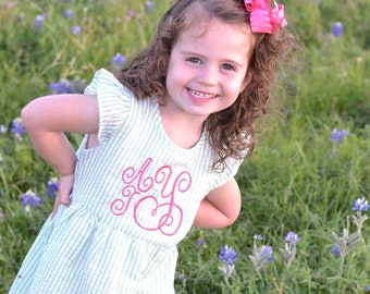 May include: A young girl with curly brown hair wears a light green and white striped dress with a pink embroidered monogram. She is standing in a field of bluebonnet flowers.