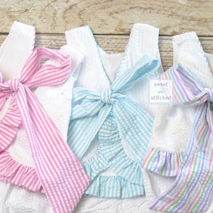 Puede incluir: Primer plano de tres tops de seersucker blancos con lazos a rayas de colores. Uno tiene un lazo rosa, otro un lazo azul claro y el tercero un lazo arco&iacute;ris pastel. El texto "sweet and stitched" es visible.