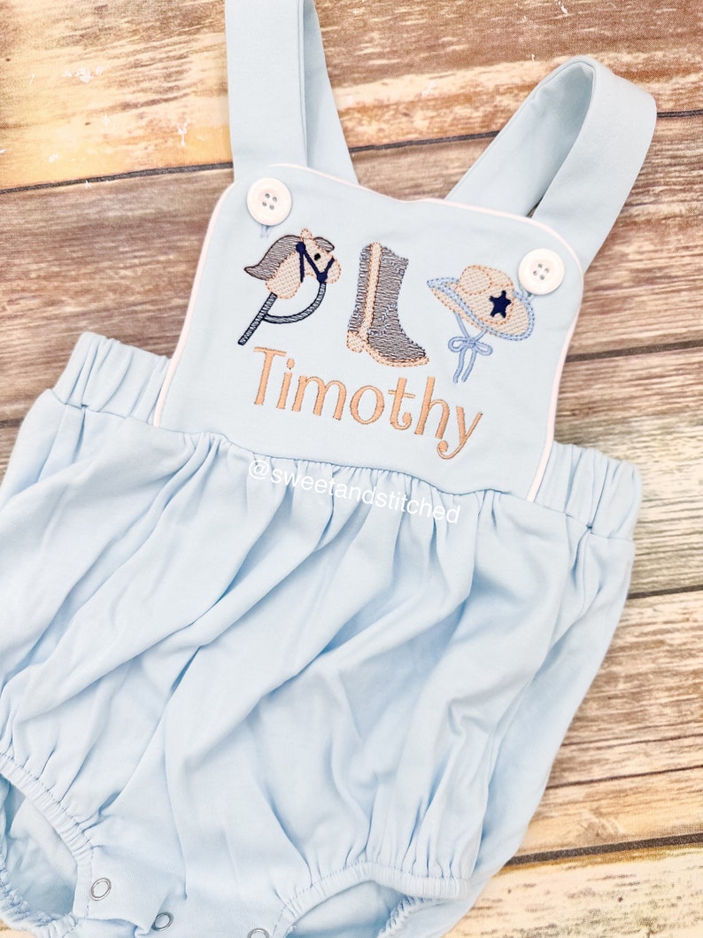 May include: Light blue baby romper with white trim and embroidered cowboy themed design. The design includes a rocking horse, cowboy boots, and a cowboy hat with a star. The name "Timothy" is embroidered below the design.