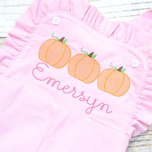 May include: Pink corduroy overalls with a ruffle trim and embroidered pumpkins. The overalls feature three orange pumpkins with green stems and the name "Emersyn" embroidered in pink cursive.