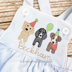 May include: Light blue baby overalls with white trim and three embroidered dogs wearing party hats. The dogs are a golden retriever, a black lab, and a black and white dog. The name "Beckham" is embroidered below the dogs. The text "@SweetAndStitched" is embroidered below the name.