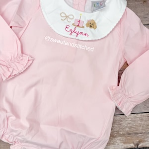 May include: A light pink baby romper with long sleeves and a white collar. The collar features embroidered details including a bow, a cake with a candle, a dog, and the name "Ezlynn". The text "@sweetandstitched" is also visible.