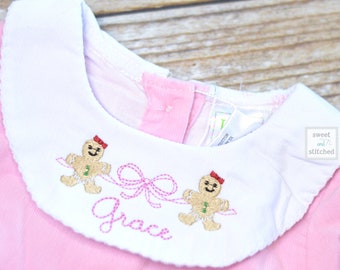 May include: Pink baby onesie with a white Peter Pan collar featuring embroidered gingerbread people and a bow. The onesie is personalized with the name "Grace".