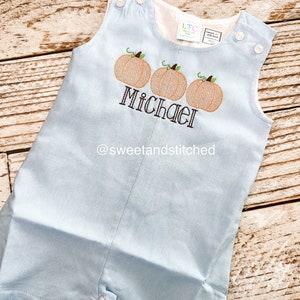 May include: Light blue baby overalls with three embroidered pumpkins and the name "Michael" in brown thread. The overalls have a white tag with the text "sweet and stitched" on the front.