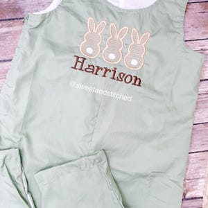 May include: Sage green baby romper with three embroidered bunny rabbits and the name "Harrison" in brown thread. The romper has white buttons and a white lining. The text "@sweetandstitched" is also visible.