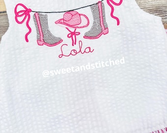 Könnte beinhalten: Weißes, ärmelloses Kleid mit rosa und weißen Streifen am Saum. Das Kleid hat ein rosa gesticktes Design mit einem Cowboyhut, Stiefeln und einer Schleife. Der Name "Lola" ist unter dem Design gestickt. Der Text "@sweetandstitched" ist unter dem Namen gestickt.