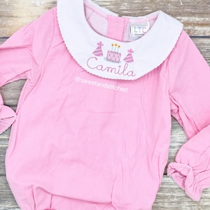 May include: Pink long-sleeve baby onesie with a white Peter Pan collar. The onesie has an embroidered birthday cake and party hats with the name "Camila" and the text "@sweetandstitched".