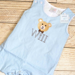 May include: Light blue sleeveless romper with a brown bear wearing a bow tie embroidered on the front. The romper has the name "Will" embroidered below the bear.
