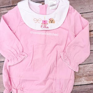 May include: Pink corduroy baby romper with a white collar. The collar features an embroidered design with a number 1, a bow, a dog, and the name "Ella". The text "@sweetandstitched" is embroidered below the collar.