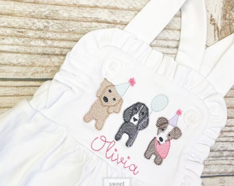 Könnte beinhalten: Weißer Strampler mit gestickten Hunden, die Partyhüte tragen und Ballons halten. Der Strampler hat einen Rüschenkragen und Träger. Der Name "Olivia" ist unter den Hunden gestickt. Der Text "sweet and stitched" ist in einem Quadrat am unteren Rand des Stramplers gestickt.