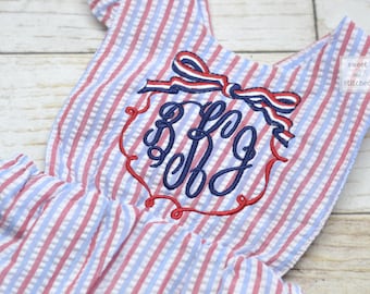 May include: A seersucker romper with red, white, and blue stripes. The romper features an embroidered monogram in a decorative frame, topped with a bow. The text "sweet and stitched" is visible in the upper right corner.