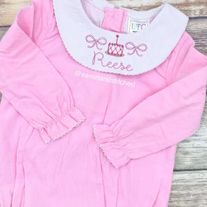 May include: Pink long-sleeve baby onesie with a white Peter Pan collar and pink embroidered bows, a birthday cake, and the name "Reese". The onesie has a scalloped trim on the sleeves and a ruffled collar. The text "@sweetandstitched" is embroidered below the name.