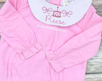 Peut inclure: Combinaison rose à manches longues pour bébé avec un col blanc Peter Pan et des nœuds brodés roses, un gâteau d'anniversaire et le nom "Reese". La combinaison a une bordure festonnée sur les manches et un col à volants. Le texte "@sweetandstitched" est brodé sous le nom.