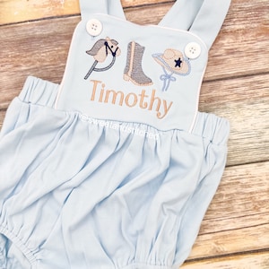May include: Light blue baby onesie with white straps and white trim. The onesie has an embroidered design of a rocking horse, cowboy boot, and cowboy hat with a star. The name "Timothy" is embroidered below the design.
